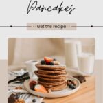 A stack of banana cocoa pancakes topped with strawberries, blueberries, and nuts sits invitingly on a plate, perfectly paired with a glass of milk.