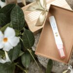 A pregnancy test showing two lines, indicating 4 weeks pregnant, is nestled inside an open gift box surrounded by green leaves and a white flower.