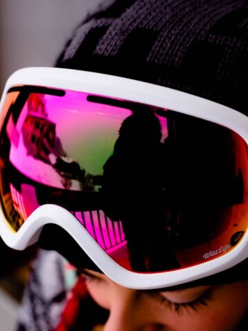 Close-up of a person wearing the best ski goggles with a black hat. The reflective goggles show the stunning reflection of snow-covered buildings and a railing.