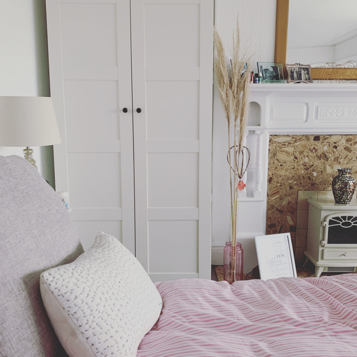 a pink striped bed with a white wardrobe and fireplace in the background. a vase of pampas grass sits beside the fireplace