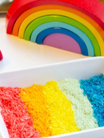 A container with coloured rice for sensory play, arranged in sections of red, yellow, green, and blue. A colorful wooden rainbow toy is in the background.