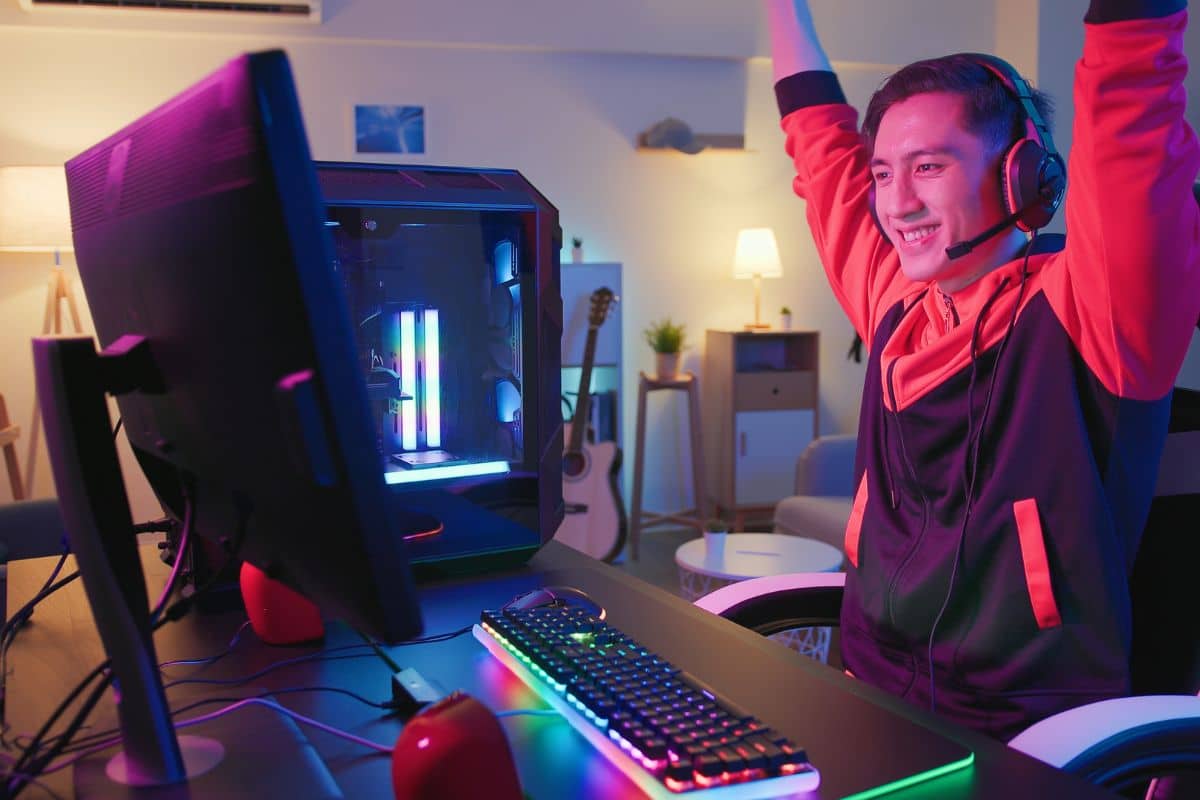 a happy man holding his hands in the air as he looks at his computer. He is wearing gaming headphones and the keyboard is neon.