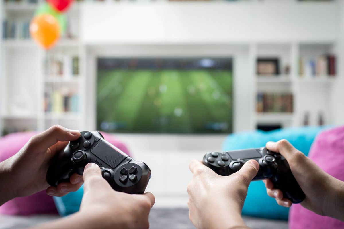 a picture showing the hands of two people holding playstation controllers and a blurry tv in the background.