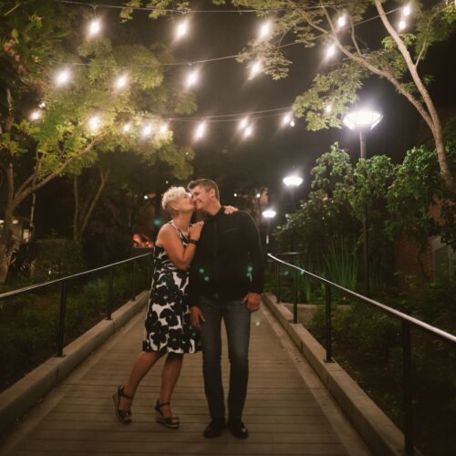 a loved up couple cuddling on a bridge with fairy lights above them.