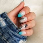 Why are acrylic nails so in fashion now? A hand with a manicure featuring turquoise and white ombre acrylic nails with black leaf-like designs, against a denim fabric and white fur background.