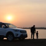 An adult and a child, considering buying a new family car, standing by a car at sunset near a body of water.
