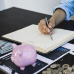 The everyday guide to taking control of your finances A person writing in a notebook with a piggy bank and money on the table indicating financial planning focused on reducing expenses or budgeting.