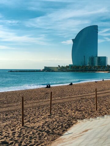 Barcelona beach with people and paddleboarders, a prominent curved blue skyscraper in the background, and clear skies.