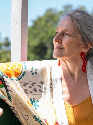 Elderly woman with gray hair, wearing a colorful blouse and earrings, leaning on a railing outdoors, looking contemplatively to the side, aging gracefully.
