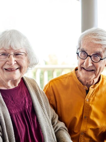 Elderly couple smiling, seated together with glasses; woman in a purple top, man in a yellow shirt enjoying quality lifestyles in a light-filled porch setting.