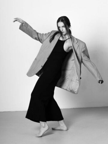 A fashion icon in a flowing pose, wearing an oversized blazer and black pants, standing barefoot on a plain background.