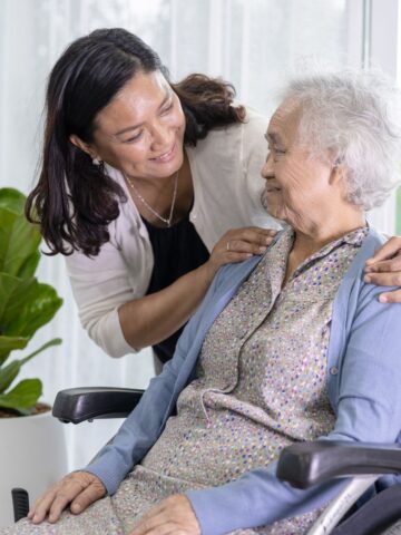 The Sandwich Generation - Balancing Care for a parent with your own family responsibilities A middle-aged Asian woman, embodying her family responsibilities, gently adjusting an elderly woman's collar as they smile at each other, the elder in a wheelchair.
