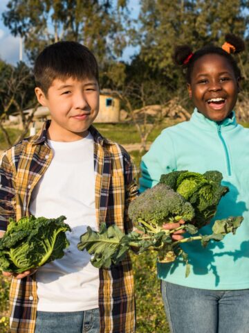 Two children smiling and holding vegetables in a garden, one Asian boy and one Black girl, enjoying the benefits of gardening with a sunny backdrop of greenery.