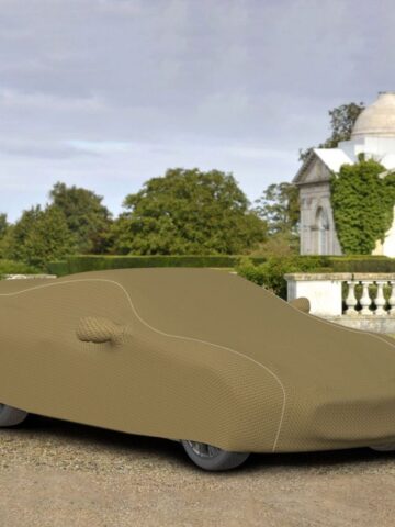 A covered car protected by a car cover parked in front of an elegant historic mansion with lush greenery and a cloudy sky in the background.