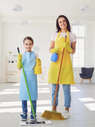 A woman and a young girl, both wearing aprons and gloves, happily holding cleaning supplies in a bright, spacious room, ready to tackle the forgotten parts during their cleaning process.