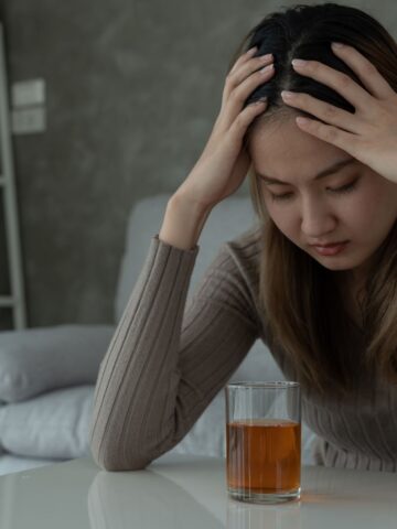 A young woman sits at a table, looking distressed with her hands on her head, with a glass of amber liquid in front of her, symbolizing her struggle with alcohol addiction.