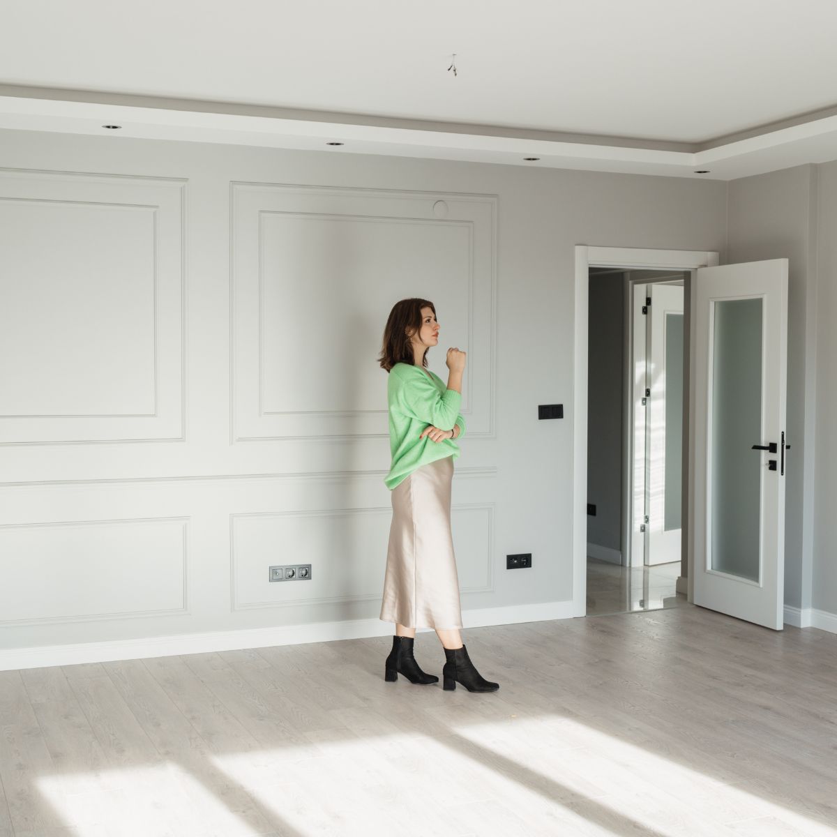 A person in a green sweater and beige skirt stands in an empty, well-lit room with white walls and light wooden floors, exploring various home options.