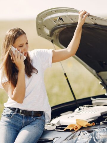 A woman in a white t-shirt and jeans is talking on the phone while leaning on a car with an open hood, ensuring satisfaction with her used car purchase. A yellow cloth is placed on the car's front.