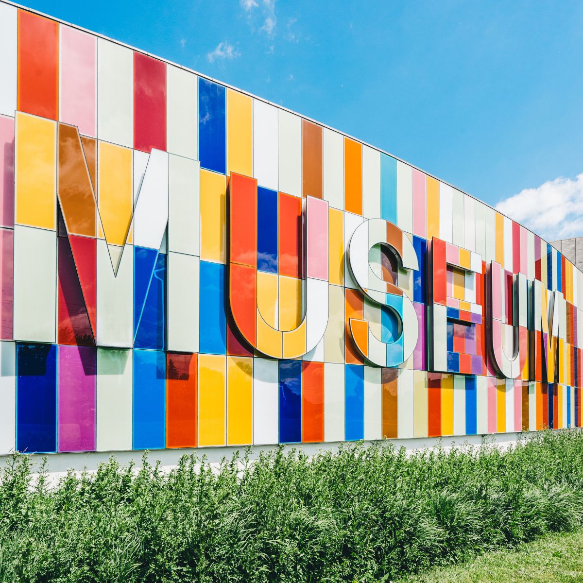 The colorful museum facade features large letters spelling "MUSEUM" against a backdrop of blue sky and green shrubs. For your next vacation, consider these travel tips: visit museums like this one to explore vibrant cultural experiences!