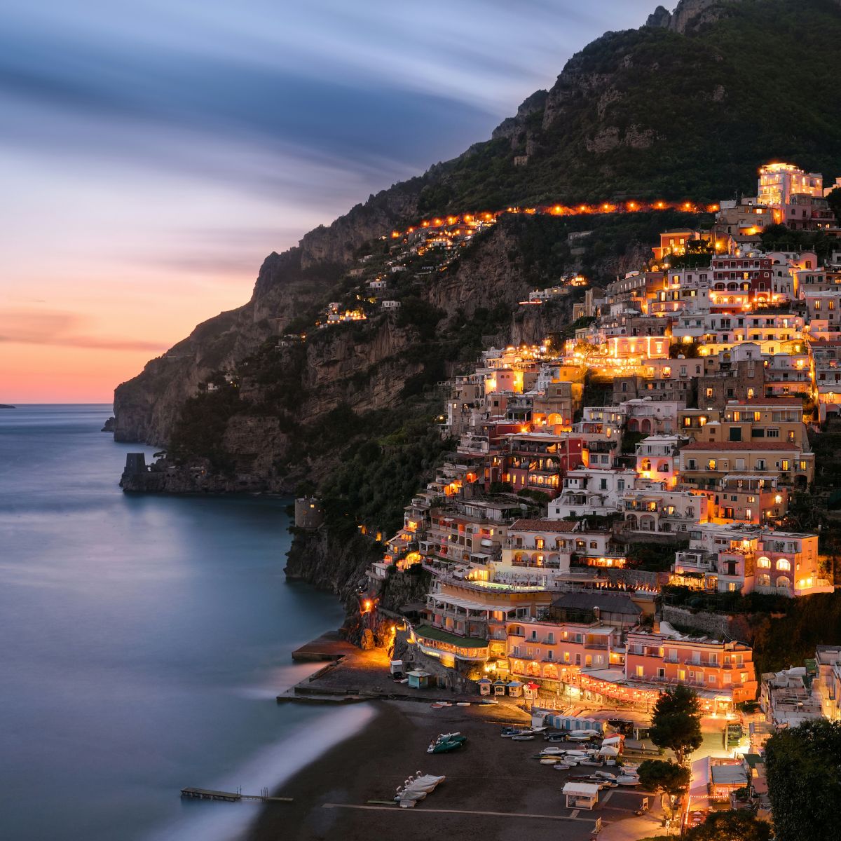 View of Positano at dusk with illuminated buildings on a steep hillside overlooking the sea, featuring a calm waterfront—a must-visit gem in Europe's stunning coastal regions.