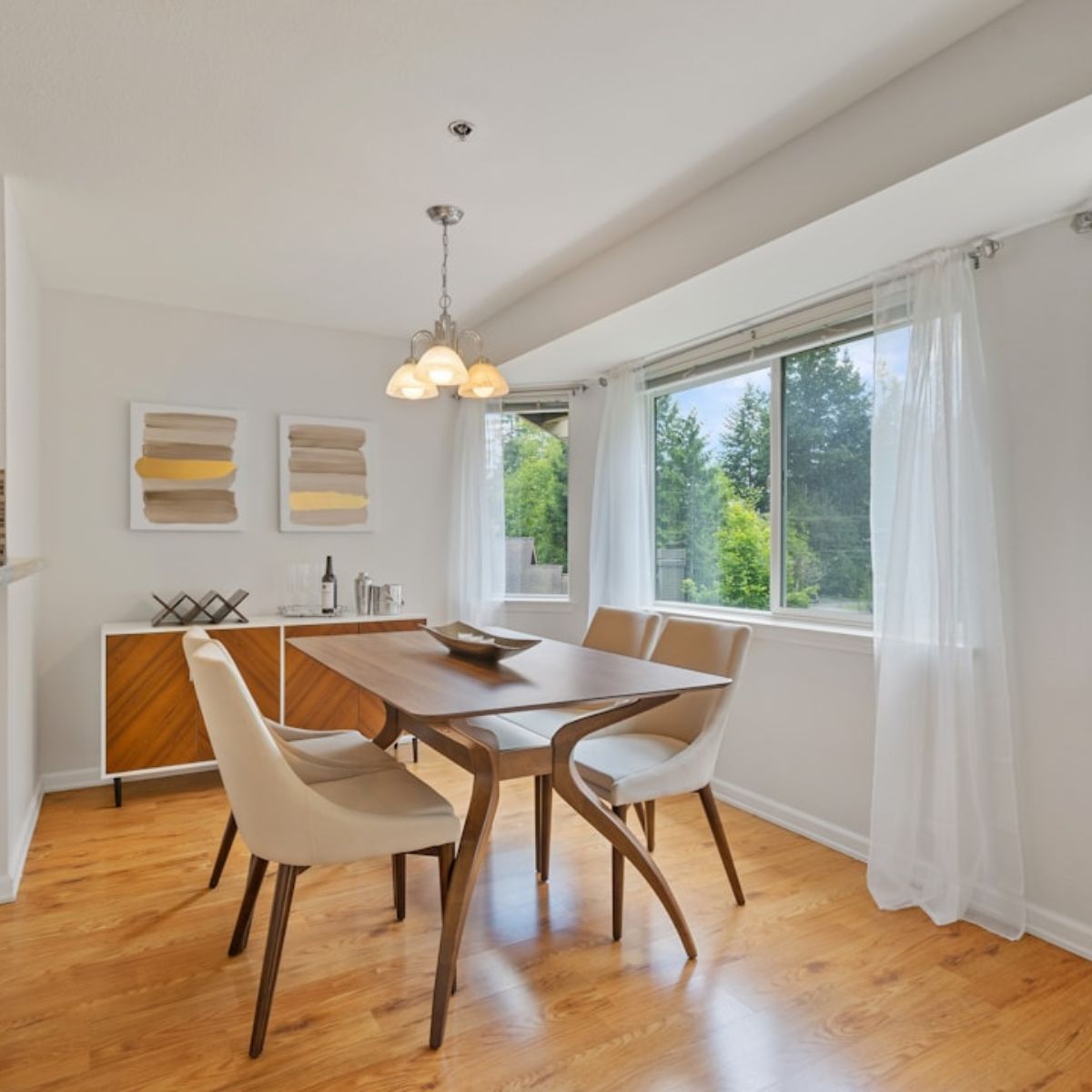 A dining area with a wooden table, four chairs, and a modern sideboard under two abstract paintings. Flooring professionally installed adds an extra touch of sophistication. Large windows with white curtains let in natural light.