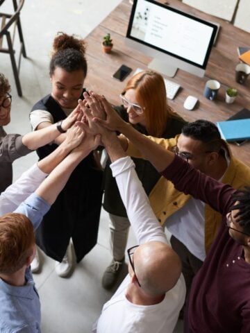 A diverse group of eight people stand in a circle, placing their hands together in the center, in a collaborative gesture at an office workspace with a computer and notebooks on the table-demonstrating top internship tips for effective teamwork.