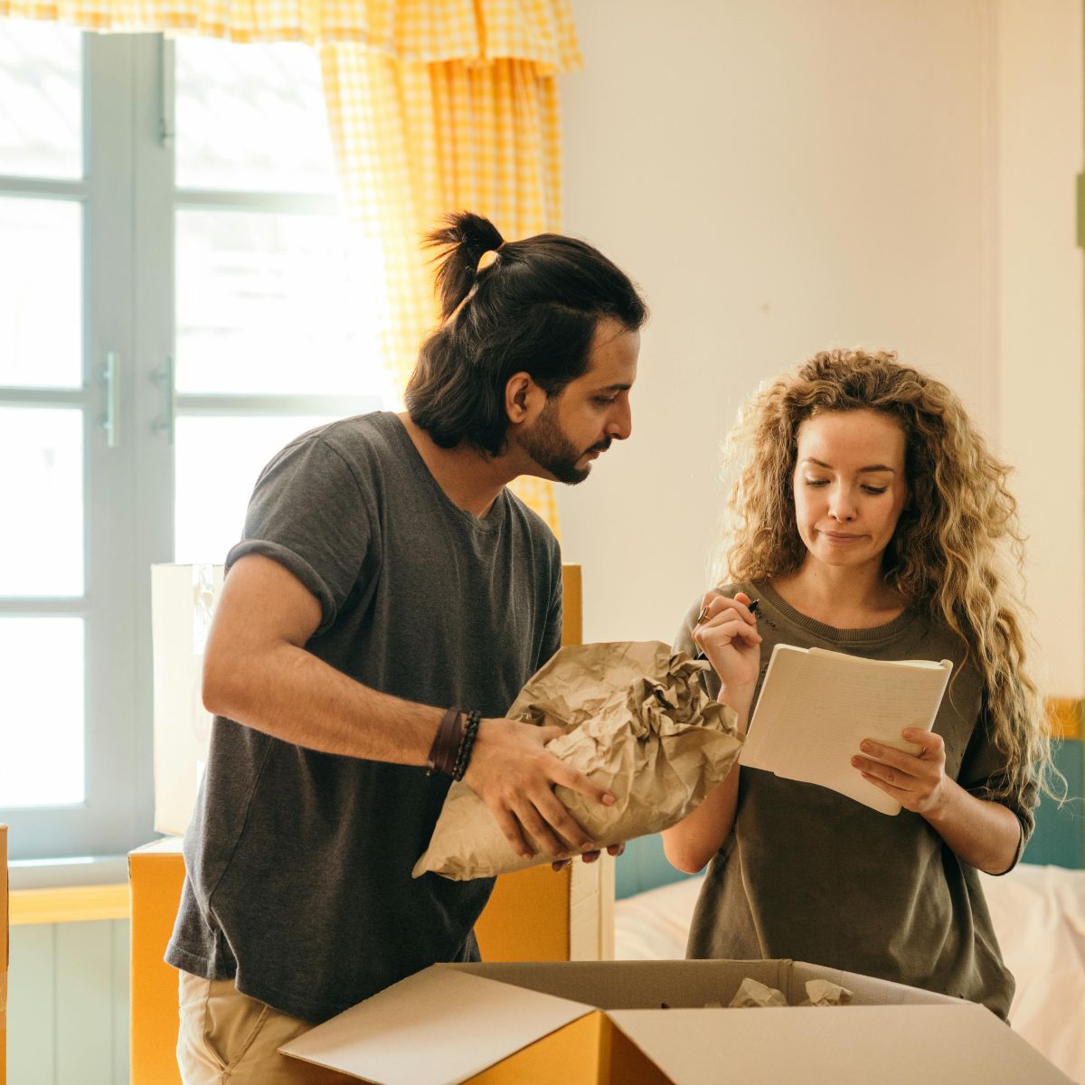 A man and woman are packing items into a box. The woman, holding a notepad and pen, checks off a list of moving tips, while the man wraps something in brown paper. They are standing in a well-lit room near a large window, making their stress-free relocation smoother and more organized.
