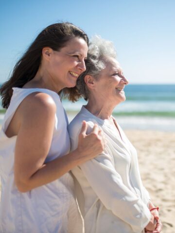 Two women, one younger and one older, smiling and standing side by side on a sunny beach with the ocean in the background, showcasing the joy that comes from caring for elderly parents.