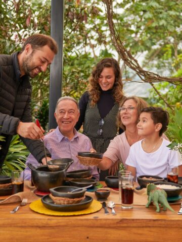 What Are The Advantages of Multi-Generational Living? A group of people sits around an outdoor table set for a meal. One person stands, serving food, while the others smile and interact, showcasing the advantages of multi-generational living. Plates, glasses, and decorations are on the table.