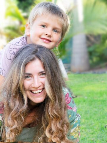 A woman smiles as a young boy sits on her back in an outdoor setting with greenery in the background, epitomizing the joy and balance possible when scholarships for mothers help them go back to school.