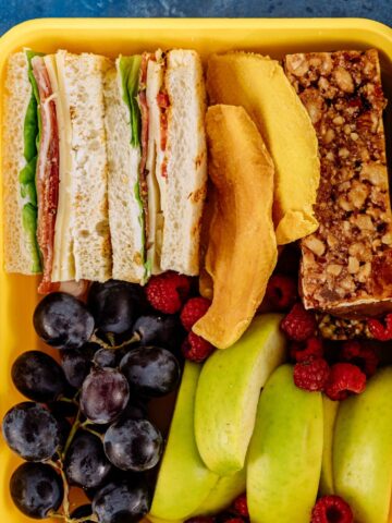 A yellow lunchbox, perfect for busy students, contains a sandwich, grapes, green apple slices, raspberries, dried mango pieces, and a granola bar on a blue surface. These mom-approved college snacks ensure a nutritious and delicious meal on the go.