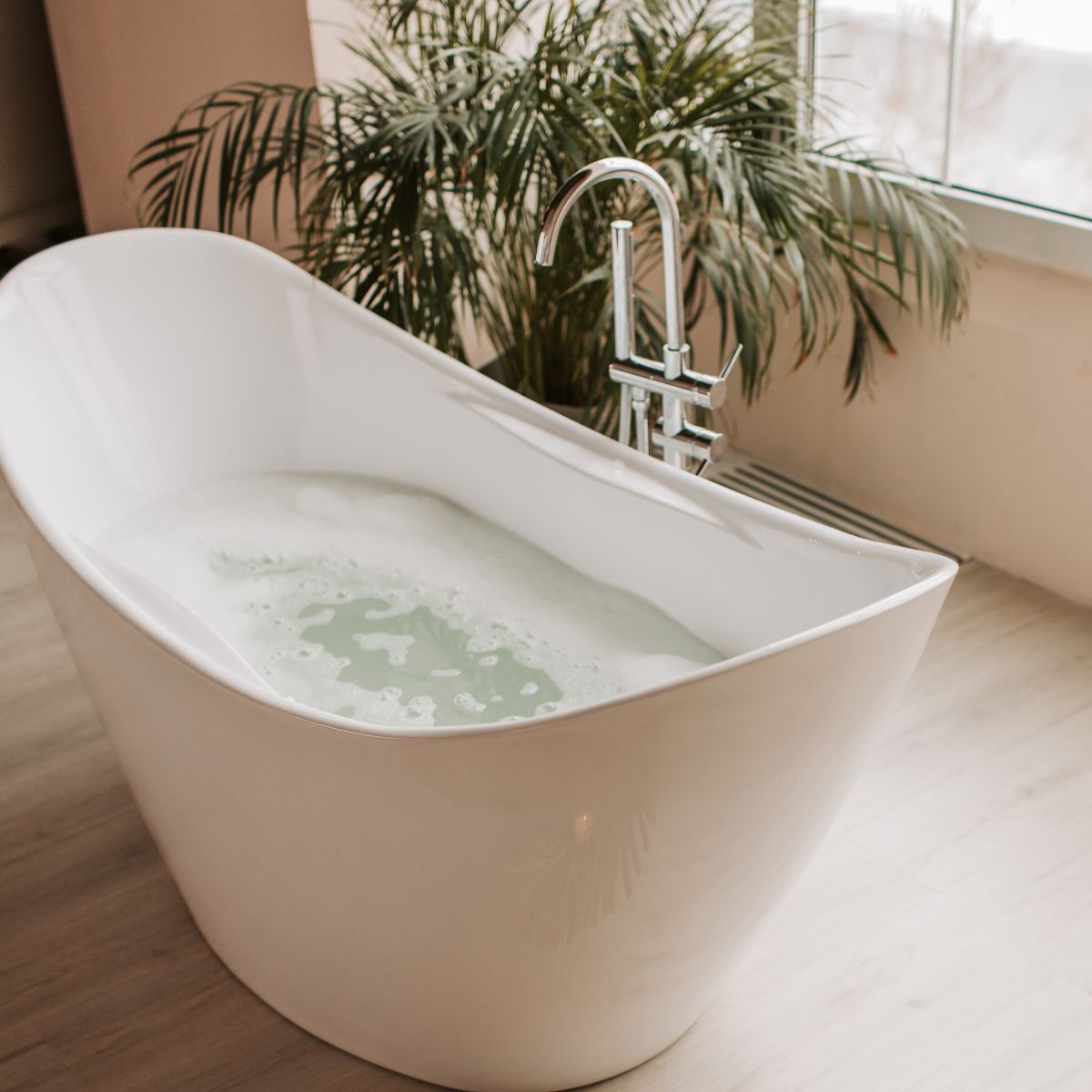 A white freestanding bathtub filled with water is placed near a window, creating the perfect bathroom with a cozy retreat vibe. A tall green plant in the background adds a touch of nature to this serene setting, making it easy to unwind and relax.