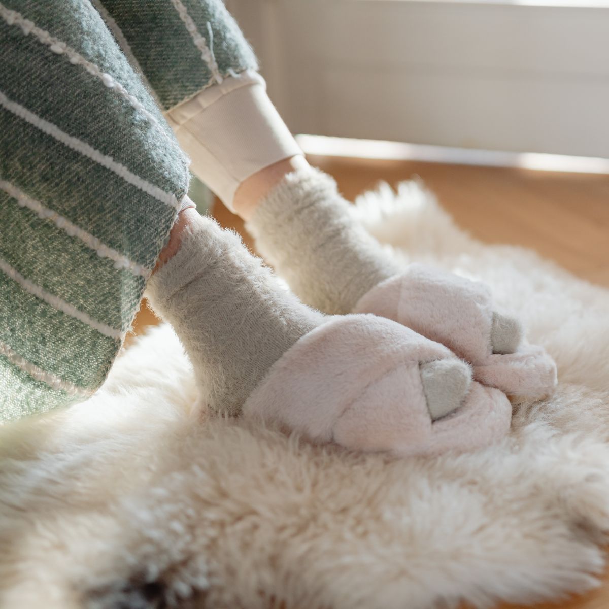 Close-up of a person wearing gray socks and fluffy slippers with a green striped blanket over their legs, seated on a fluffy rug, showcasing the relaxed etiquette of wearing shoes indoors.