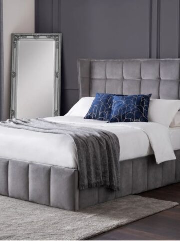 A neatly made divan bed with a tufted gray headboard and matching base, white bedding, and a gray throw blanket. Blue throw pillows are on the bed, a tall mirror and gray curtains are nearby.