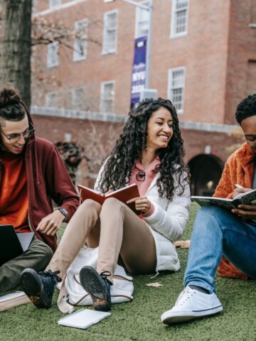 Three high school students are sitting on grass near a building, reading and smiling. Two have laptops and one has a notebook. One student, referring to an ultimate guide for college prep, points at the notebook while the others look on, laughing.