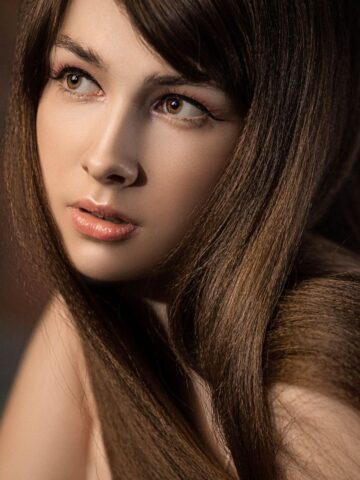 A young woman with long brown hair and light makeup looks off to the side. Despite potential hair frizz, her locks remain smooth, perhaps thanks to a conditioner enriched with fleeceflower root extract. The background is blurred.