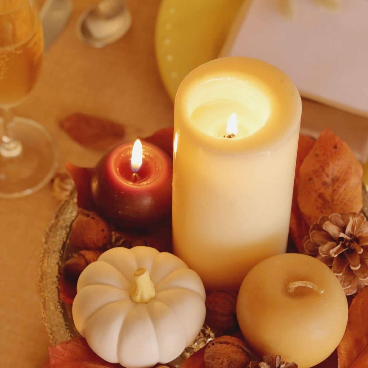 A Pinterest-inspired tabletop arrangement with a lit pillar candle, a red candle, a white pumpkin, pinecones, and autumn leaves brings cozy autumn comfort to your beige surface.