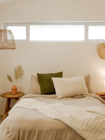 A neatly made bed with beige and green pillows, two wooden nightstands with lamps, a woven pendant light, and decorative items against a white wall with a rectangular window above creates Pinterest-inspired autumn comfort.