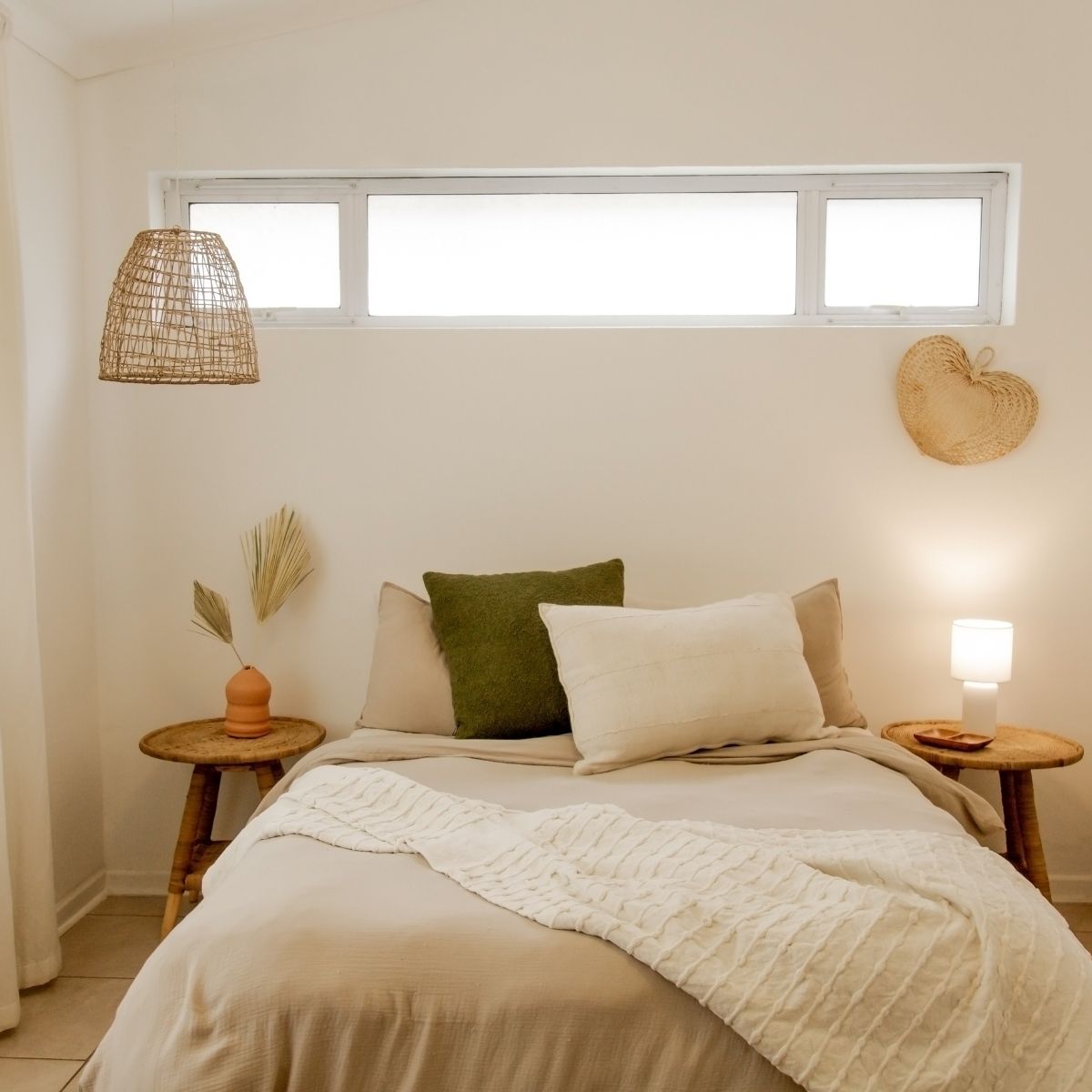 A neatly made bed with beige and green pillows, two wooden nightstands with lamps, a woven pendant light, and decorative items against a white wall with a rectangular window above creates Pinterest-inspired autumn comfort.
