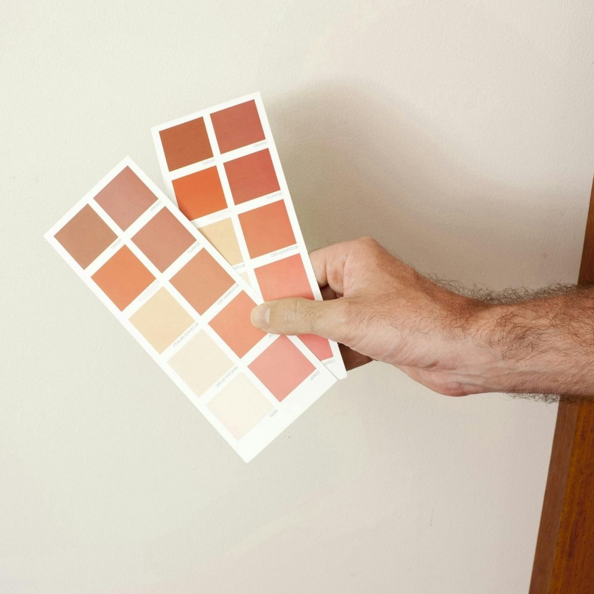 A hand holds two paint color sample cards with various shades of beige, peach, and brown—perfect autumnal colours to inspire your wardrobe or fashion choices—against a light-colored wall.