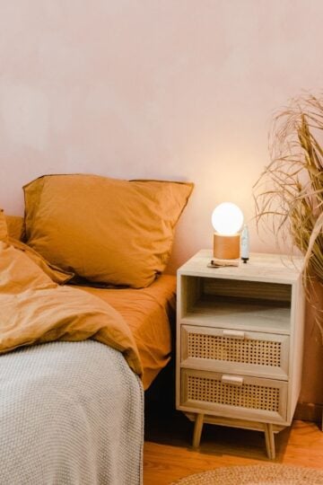 A neatly made bed with tan bedding, embracing Pinterest-Inspired autumn comfort, sits beside a nightstand with a glowing lamp, a bottle, and a vase of dried plants against a light pink wall.