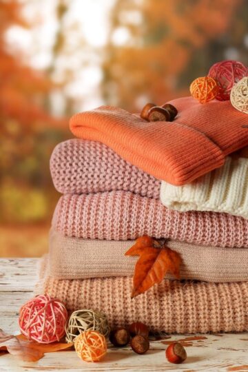 Stack of folded knit sweaters in autumnal colours on a wooden surface, perfect for your wardrobe, decorated with dry leaves, acorns, and decorative balls, with a blurred fall foliage background.