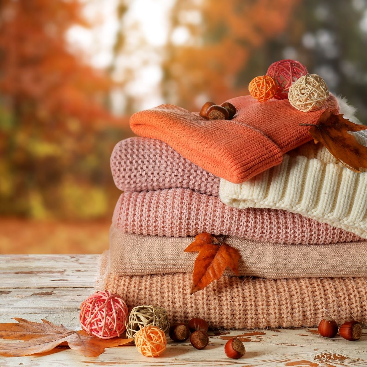 Stack of folded knit sweaters in autumnal colours on a wooden surface, perfect for your wardrobe, decorated with dry leaves, acorns, and decorative balls, with a blurred fall foliage background.