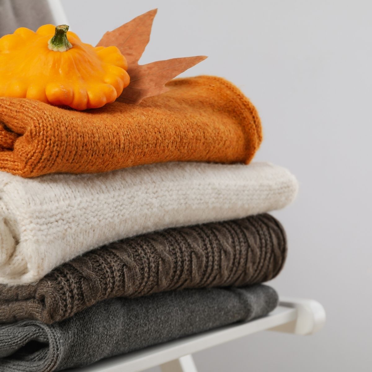 Four folded sweaters in autumnal colours—gray, brown, cream, and orange—stacked on a chair, with a small pumpkin and two brown leaves placed on top; a cozy fashion statement for your seasonal wardrobe.
