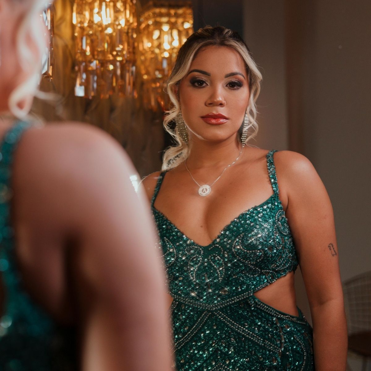 A woman in a green, sparkly dress with cutout sides stands in front of a mirror, admiring her reflection. The scene, accented by a gold chandelier, highlights her fashion-forward wardrobe.