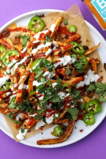A tantalizing plate of Volcano Fries, loaded with jalapeño slices, chopped cilantro, cheese sauce, sour cream, and hot sauce. Perfectly placed on a sheet of parchment paper atop a white plate, the dish invites you to customize your toppings with the seasoning bottle next to it.