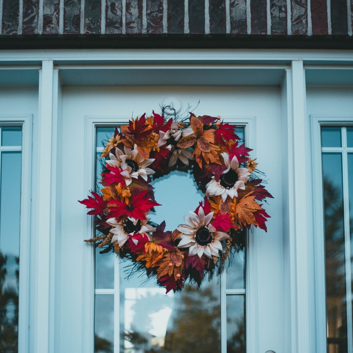A wreath made of red, orange, and yellow autumn leaves with artificial sunflowers hangs on a glass-paneled front door—an inviting touch for selling your home in a competitive market.
