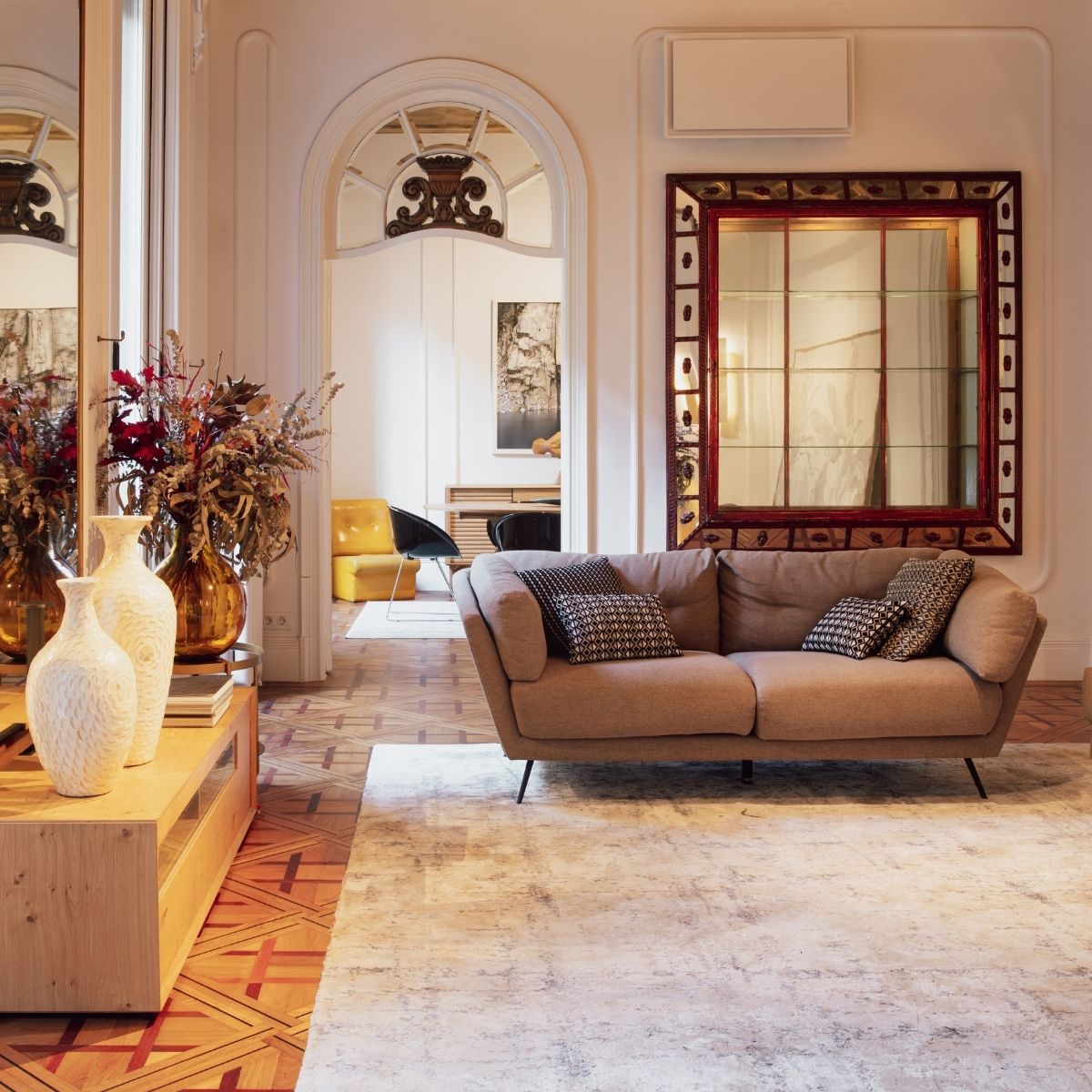 A modern living room with a beige sofa, patterned pillows, a large ornate mirror, decorative vases, and a neutral rug on a wood-patterned floor creates inviting appeal—perfect for selling your home in today’s competitive market.