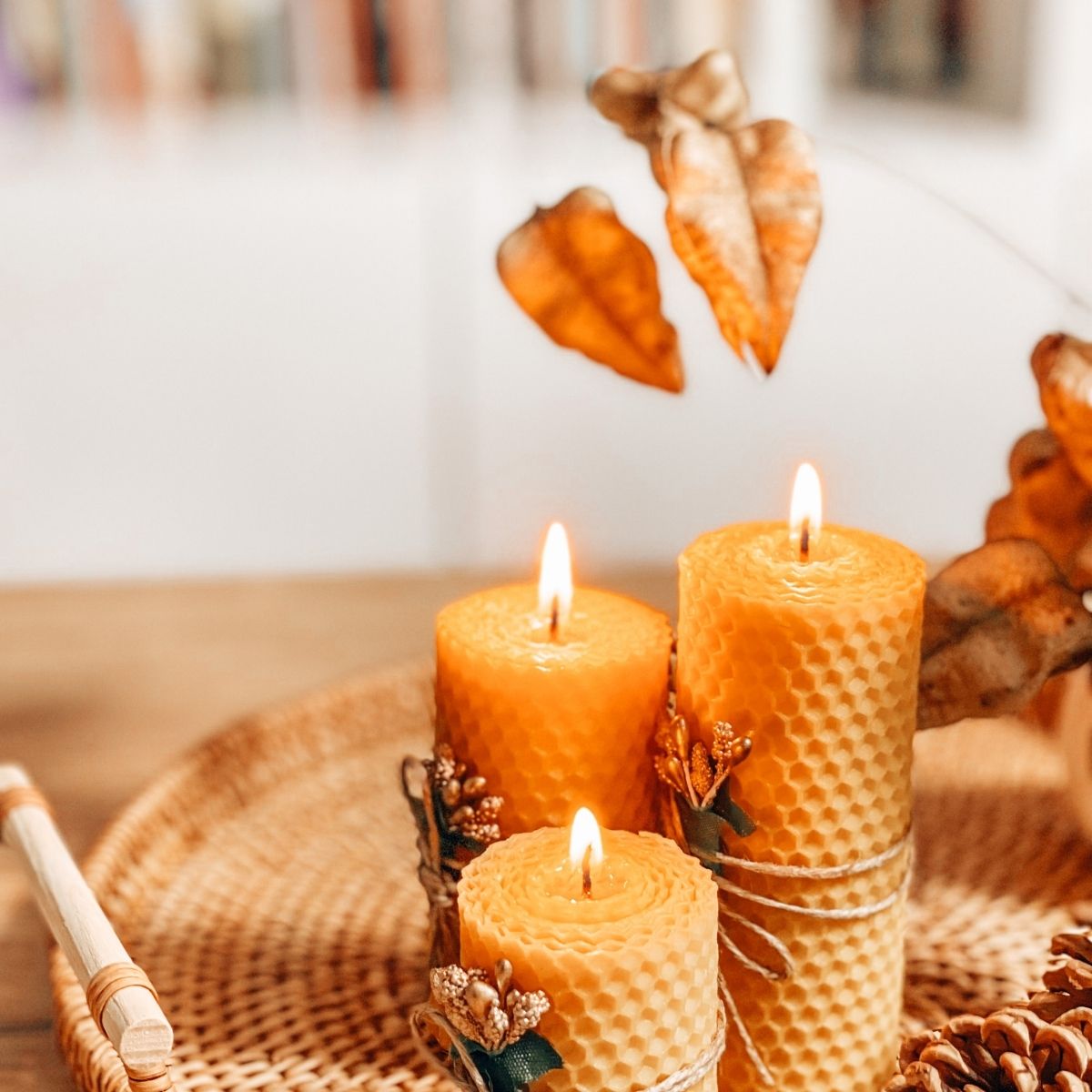 Three lit beeswax candles decorated with small sprigs and twine sit on a woven tray beside dried leaves, creating a cozy autumn ambience—perfect for selling your home in a competitive market.