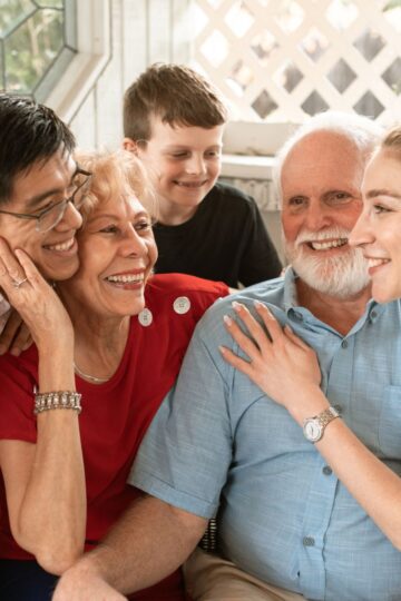 A group of five people, balancing life with joy, gathers indoors: three adults and two children. Two are seated while the others stand around them, smiling and engaging warmly, a testament to the harmony between life's different stages.